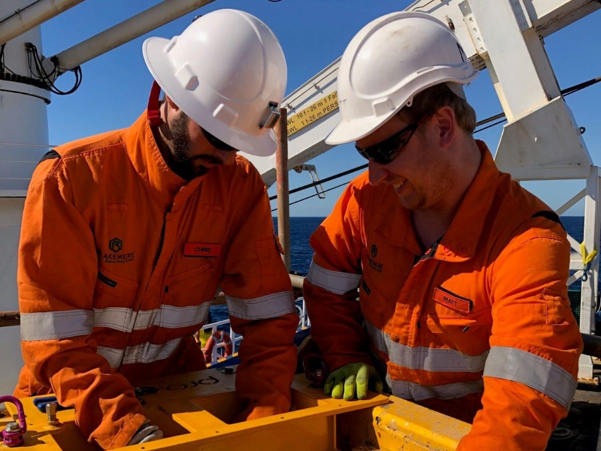Two Blakemere engineers working on a yellow tool frame on a vessel deck, the sea behind them