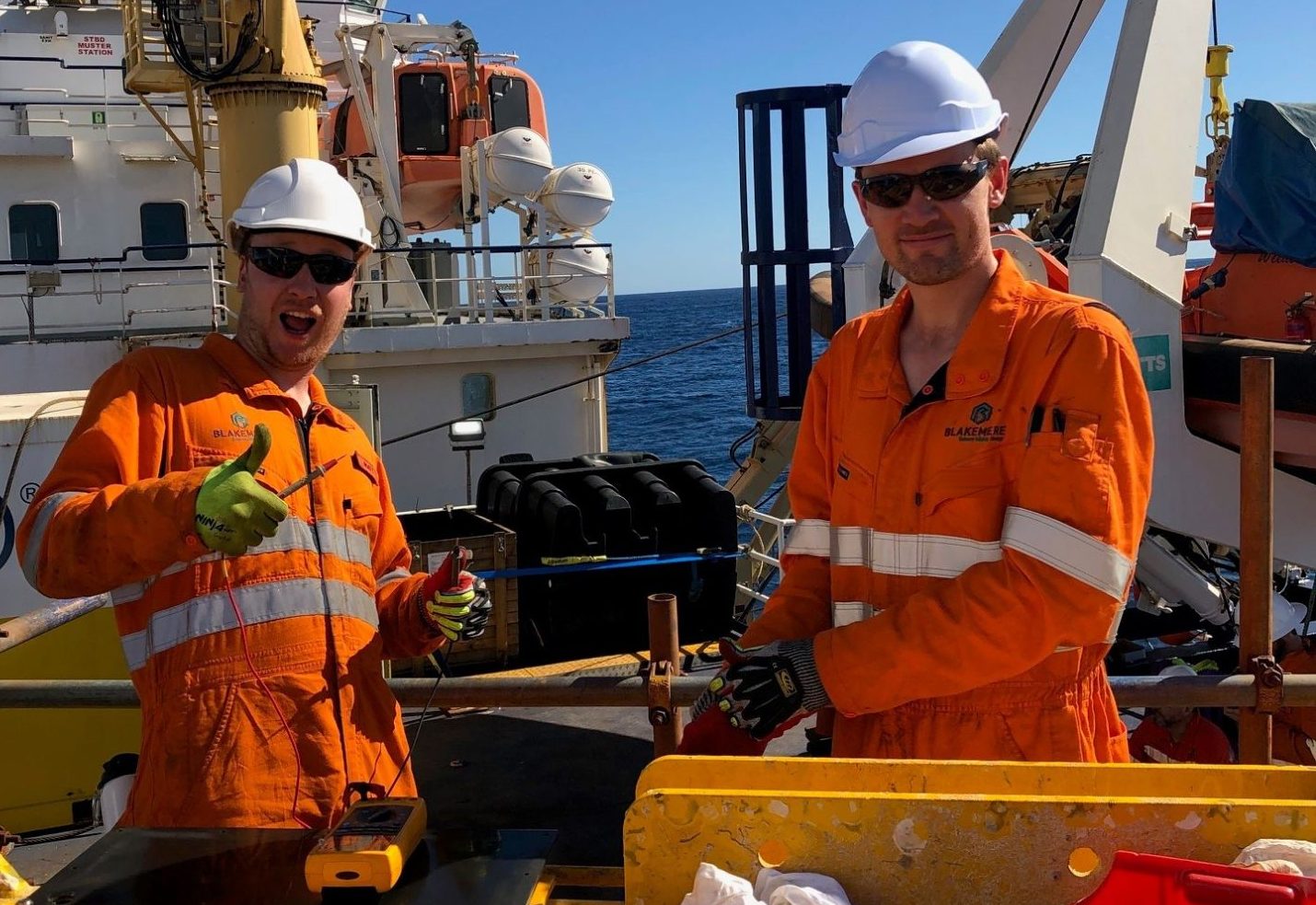 Two Blakemere technicians in branded coveralls at a tool frame on a vessel deck, one giving a thumbs up