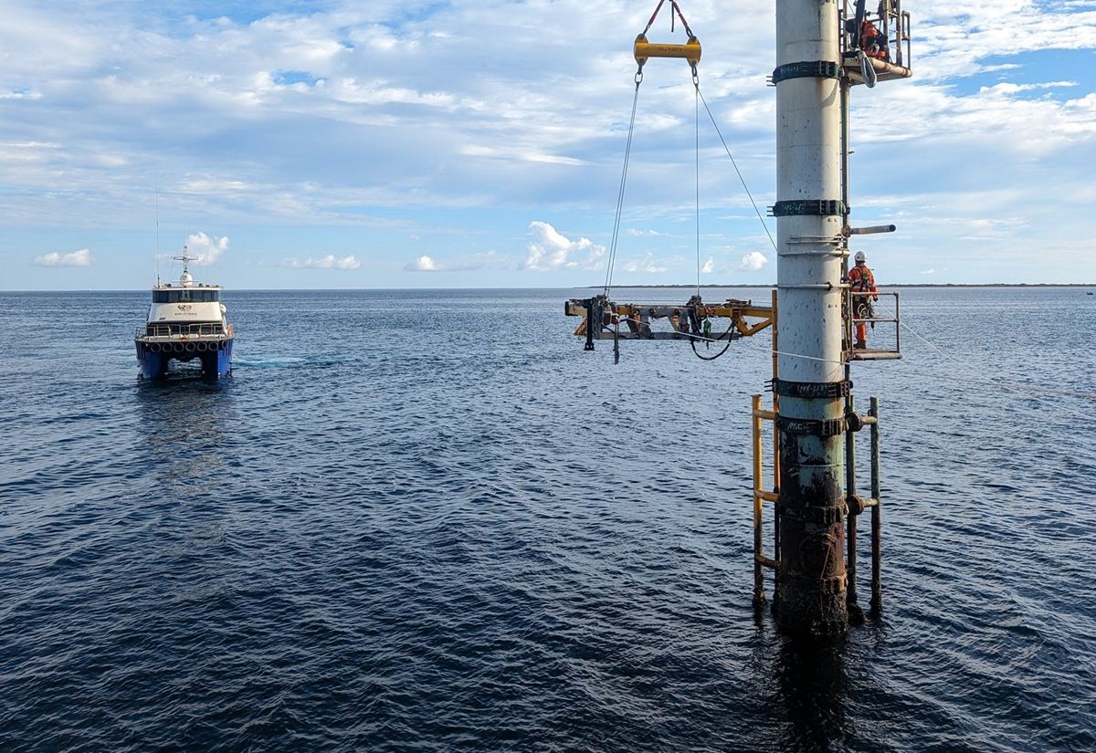 A cutting frame on the crane wire being positioned against a monopod, with technicians working from the structure and a crew boat standing off