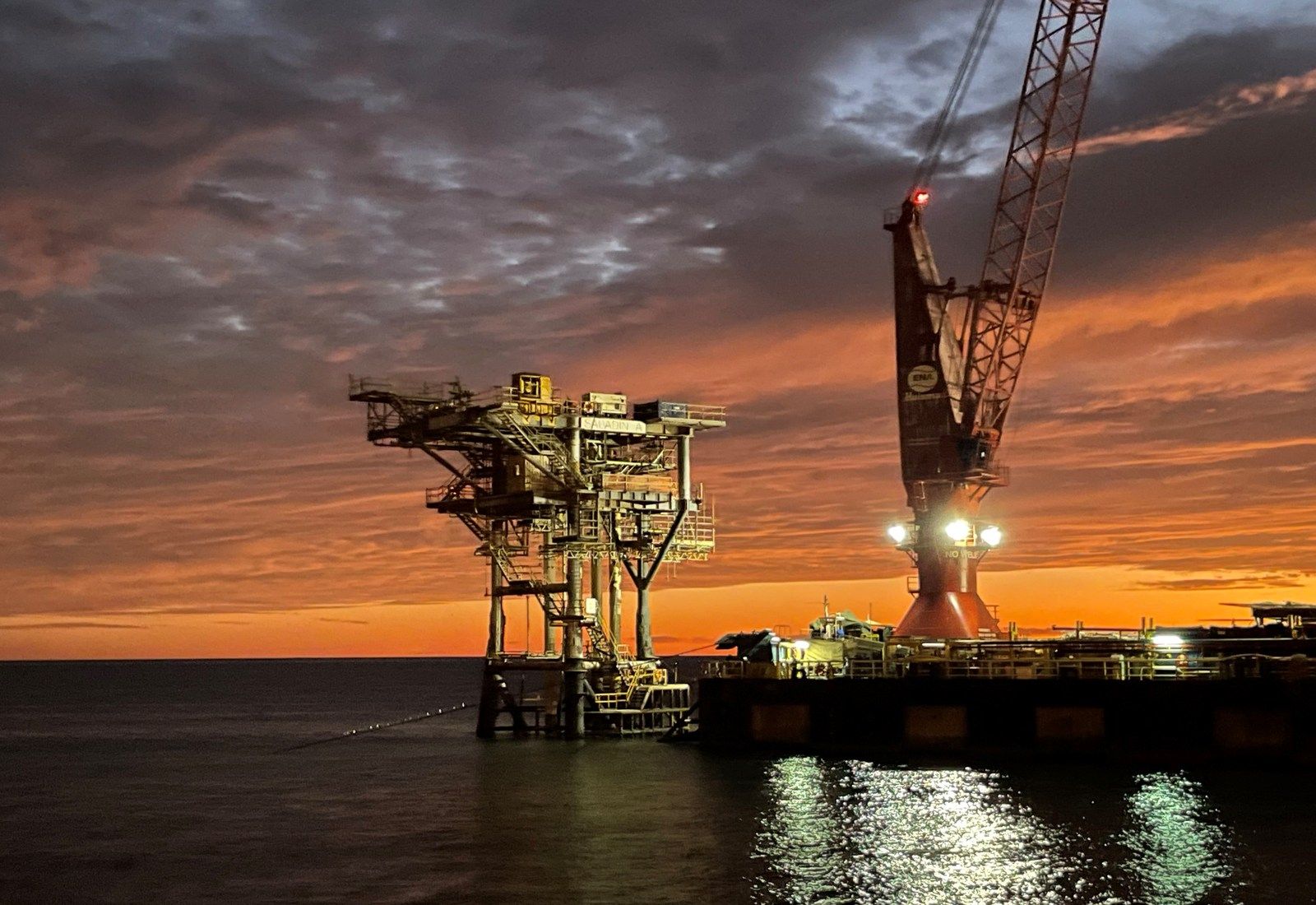 An unmanned platform alongside a heavy-lift crane barge at sunset during a removal campaign