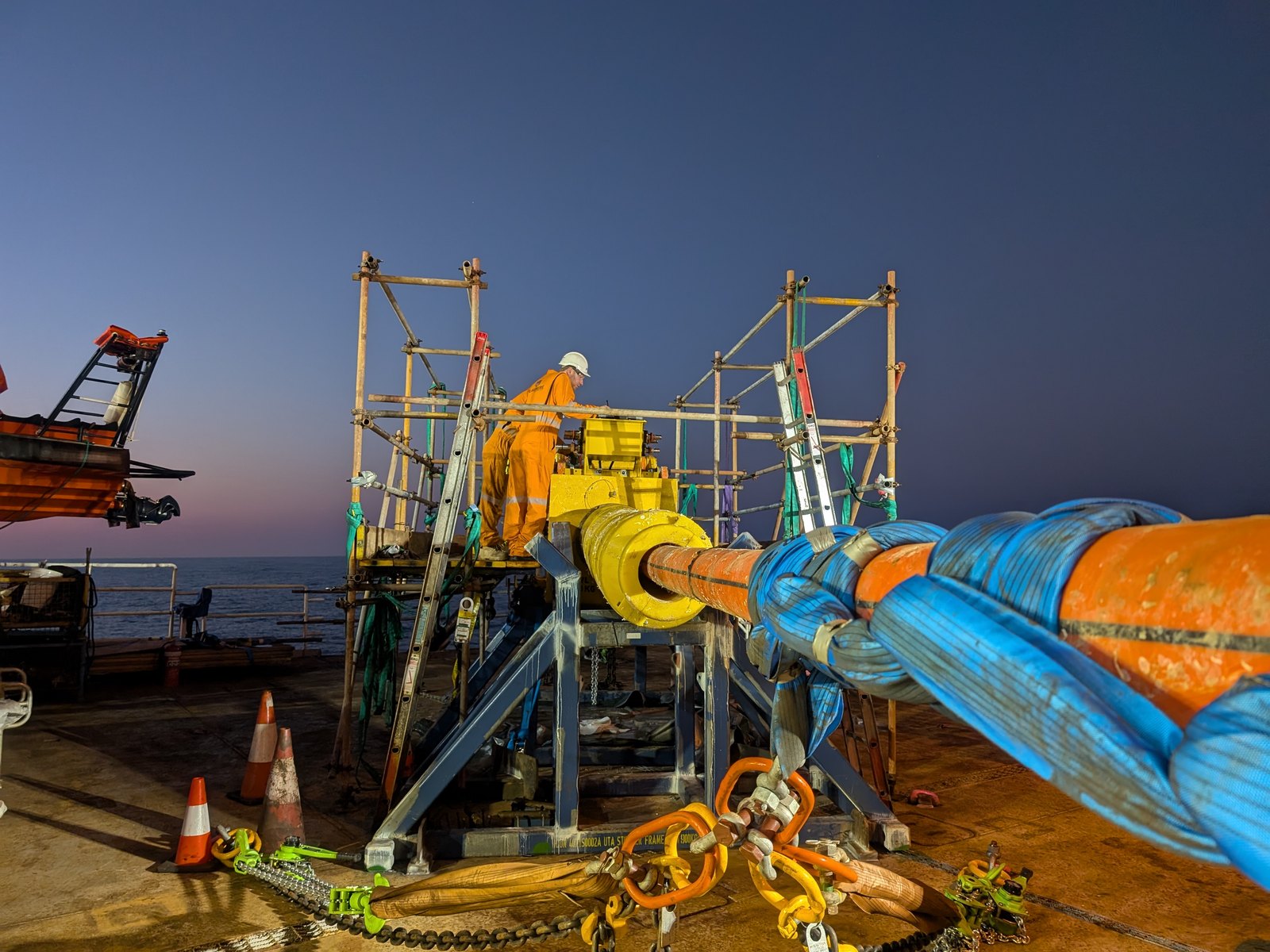 A subsea umbilical termination assembly on its stinger frame on a vessel back deck at dusk, scaffolded for access, with a Blakemere technician working at the termination head and the umbilical running off the deck