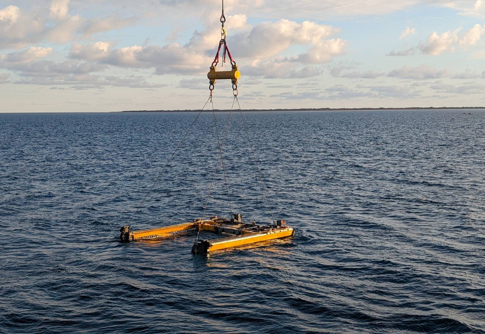 A yellow subsea cutting frame on a crane spreader bar being lowered into the sea, a low coastline on the horizon