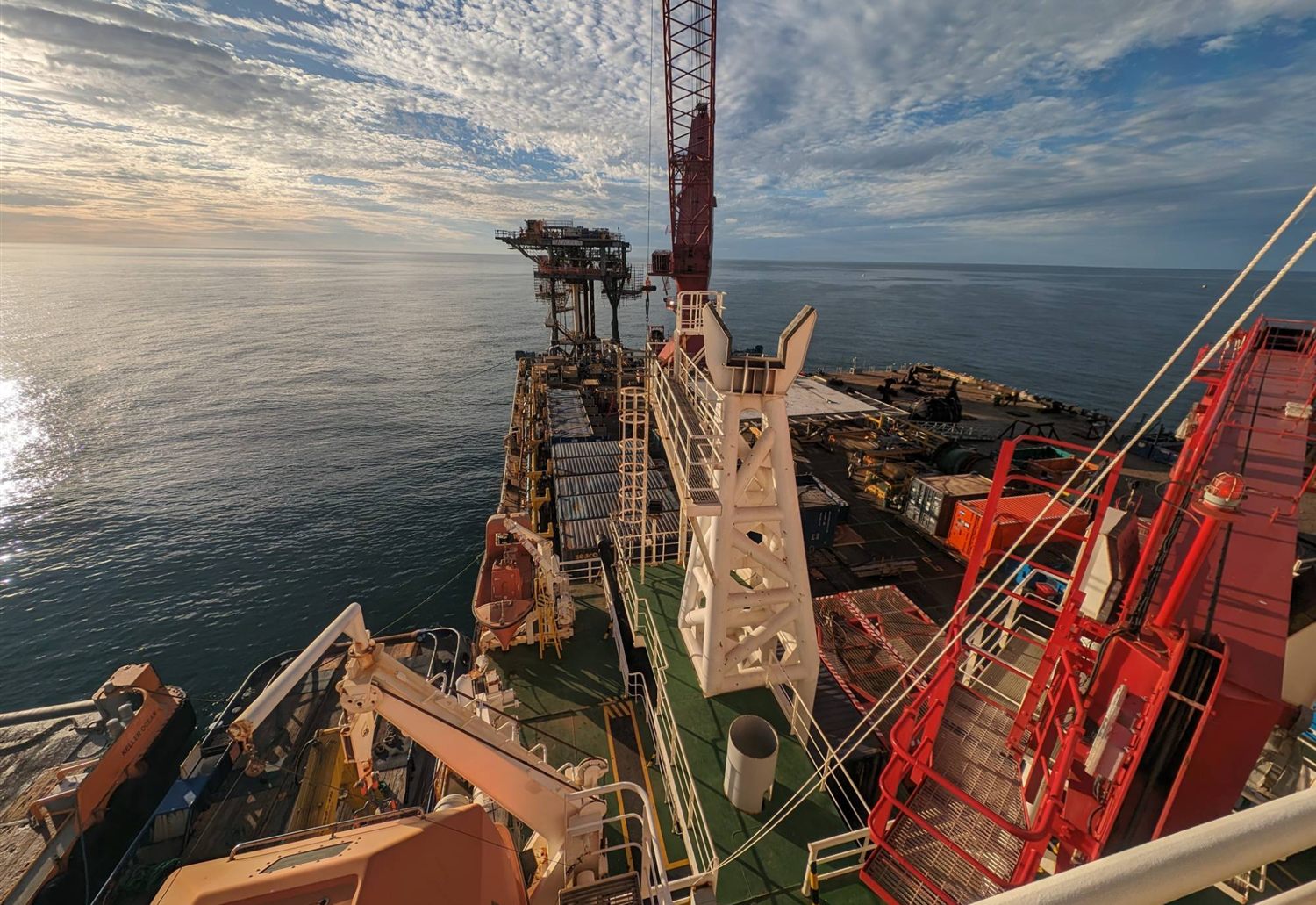 Looking along the deck of a construction vessel towards an offshore platform, with the main crane, containers and campaign equipment on deck and a support vessel alongside