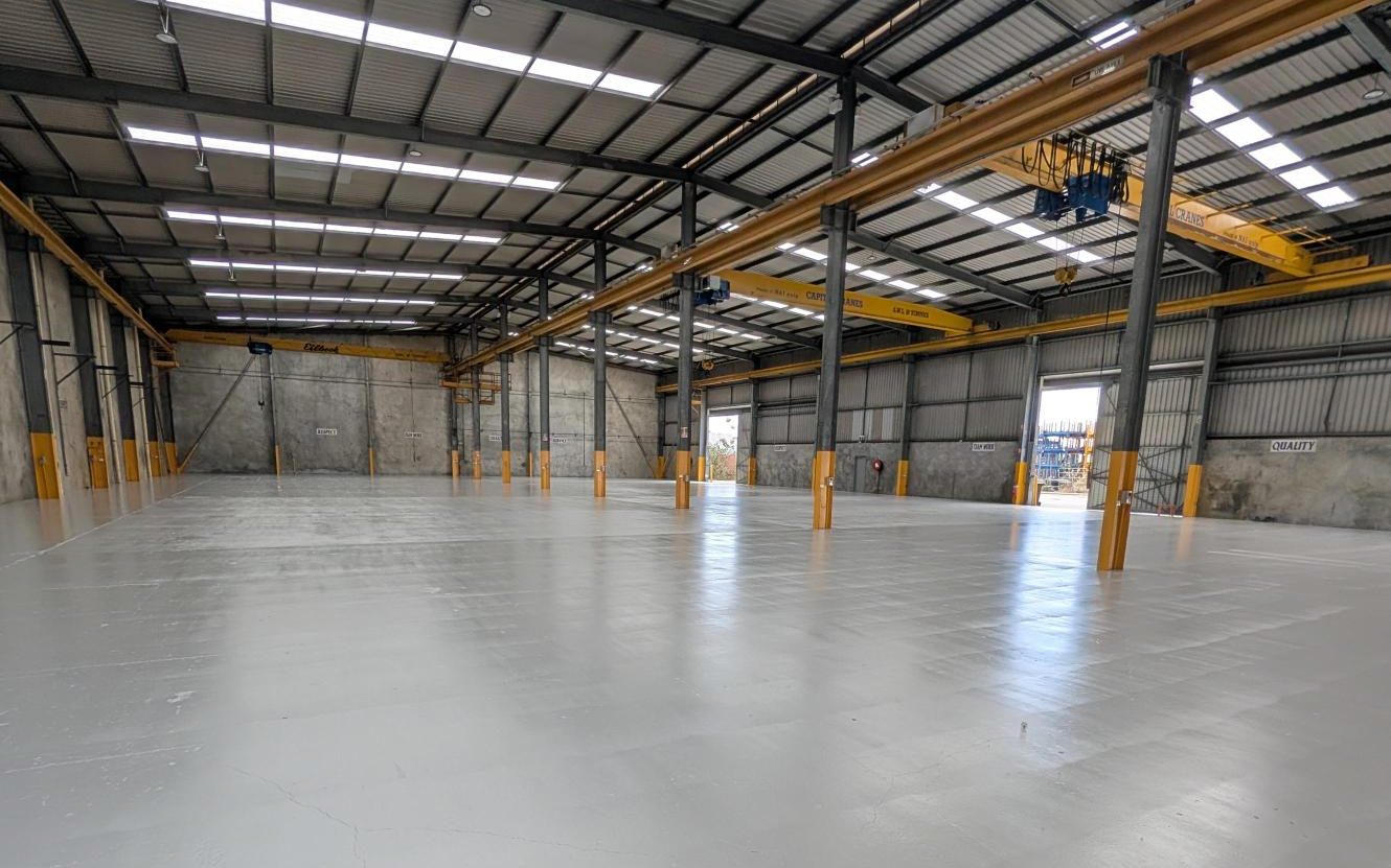 An empty, freshly coated workshop bay under skylights, with yellow overhead cranes on runway beams and a roller door open to the yard at the far end