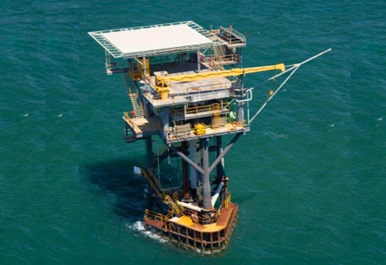 An unmanned tripod platform seen from the air, three legs carrying a multi-level topside with a crane and boat landing
